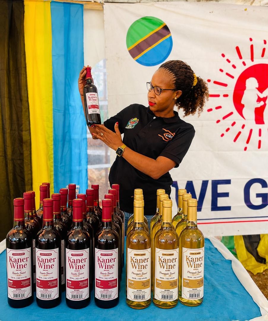 A person presenting a Kaner Wine bottle beside a display of Roselle and Pineapple bottles